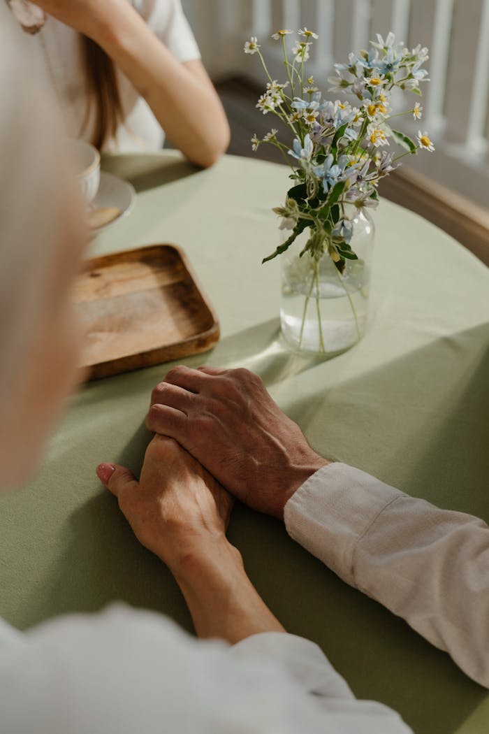 Two people holding hands across a table with a vase of chamomile, under soft daylight.